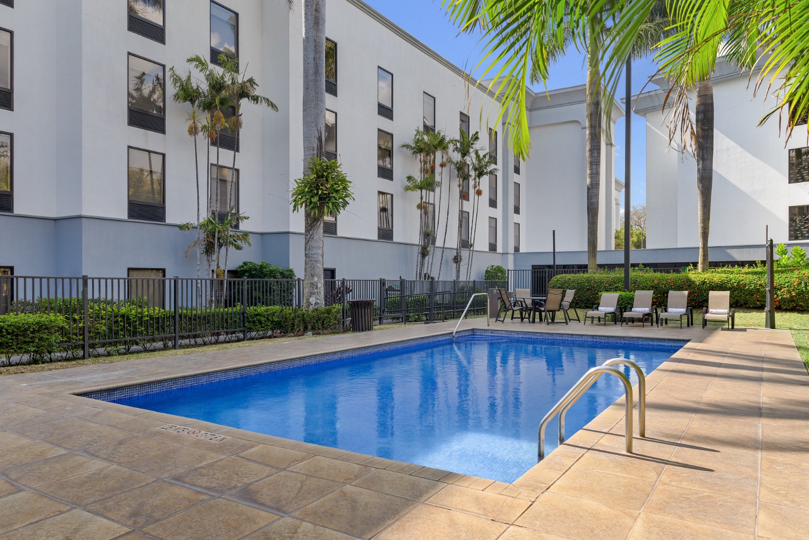 Piscina al aire libre con tumbonas, palmeras y el edificio de cuatro plantas del hotel al fondo