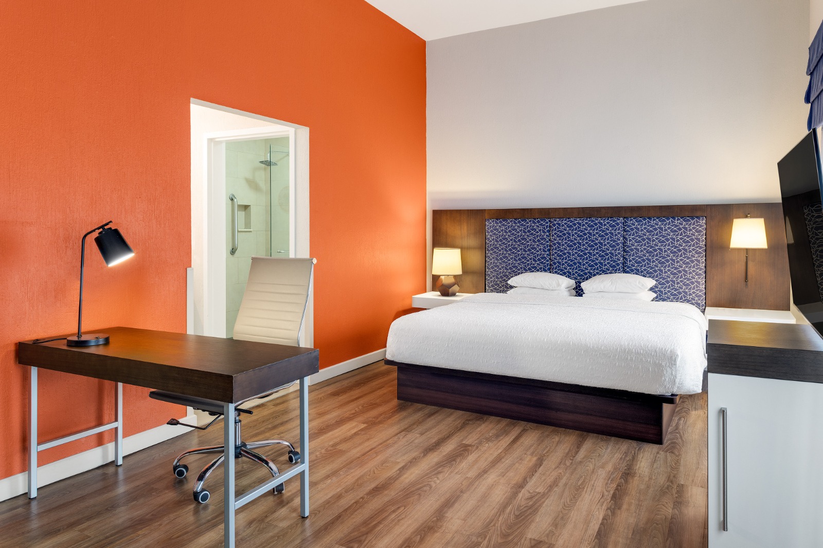 Habitación con cama king, pared naranja, escritorio de trabajo y baño con ducha de vidrio